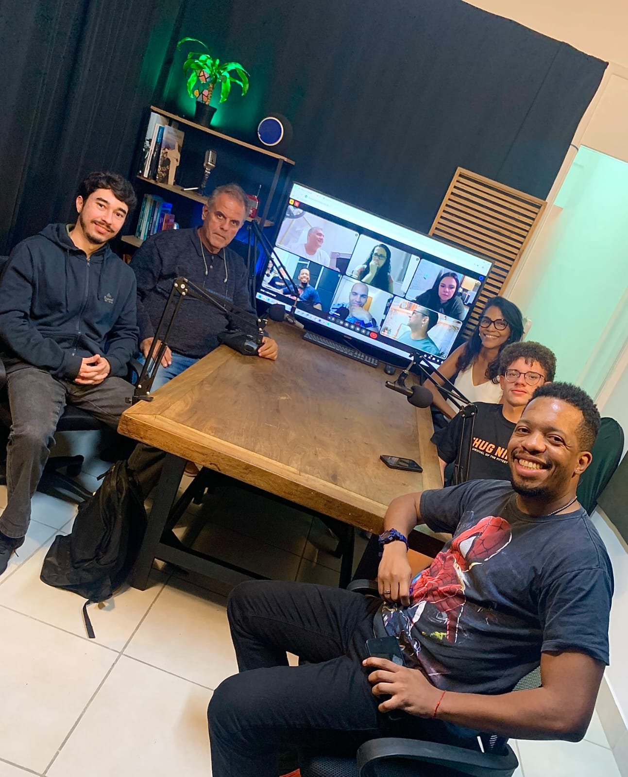 Alunos na mesa do estúdio da Escola de Rádio, com os colegas da turma online na tela ao fundo