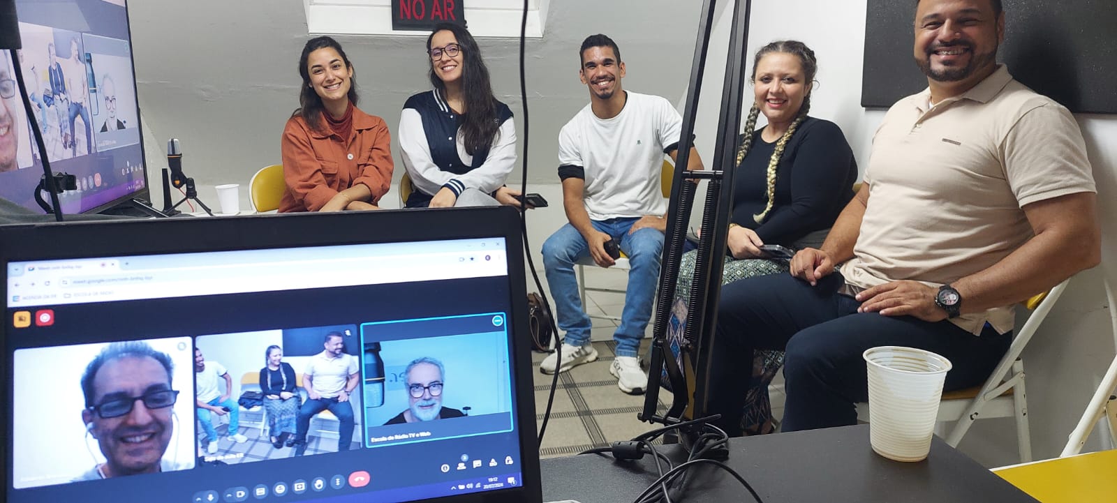 Cinco alunos no estúdio com placa NO AR e colega em chamada ao vivo no laptop