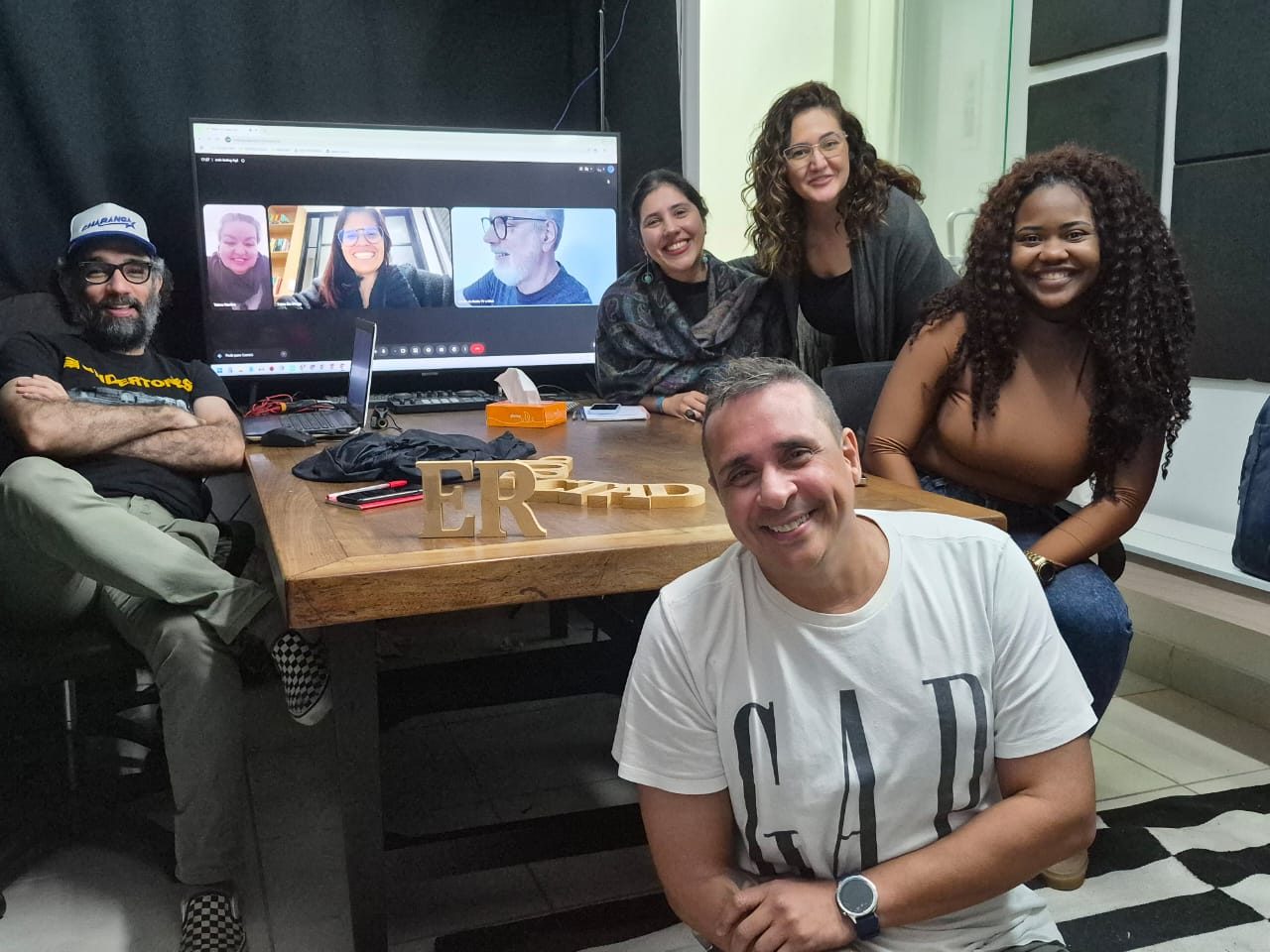 Turma ao redor da mesa com as letras ER de madeira e a aula online projetada na tela