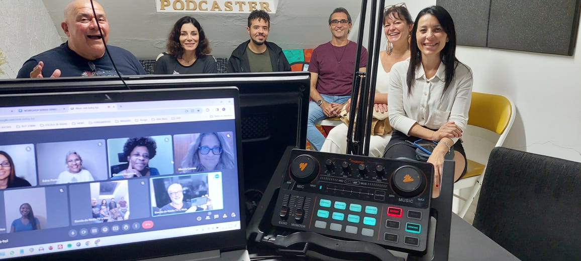 Turma na sala de podcast com parede decorada e mesa controladora no estúdio